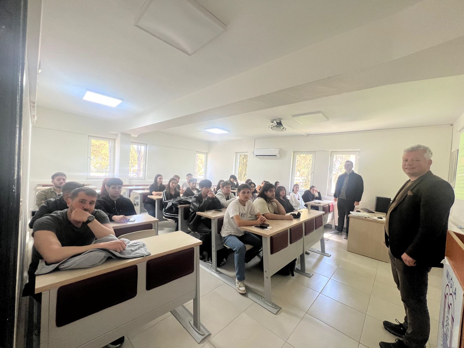 Bir derslikte yapılan toplantıda öğrenciler sıralarda oturarak kameraya doğru bakıyor. Ön planda bir konuşmacı, arka tarafta ise diğer konuşmacı öğrencilerin yanında ayakta duruyor; sınıfta projeksiyon perdesi ve geniş pencereler yer alıyor.